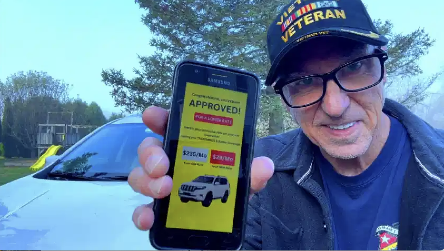 Veteran with VETERAN cap holding smartphone showing auto insurance approval with lower rates